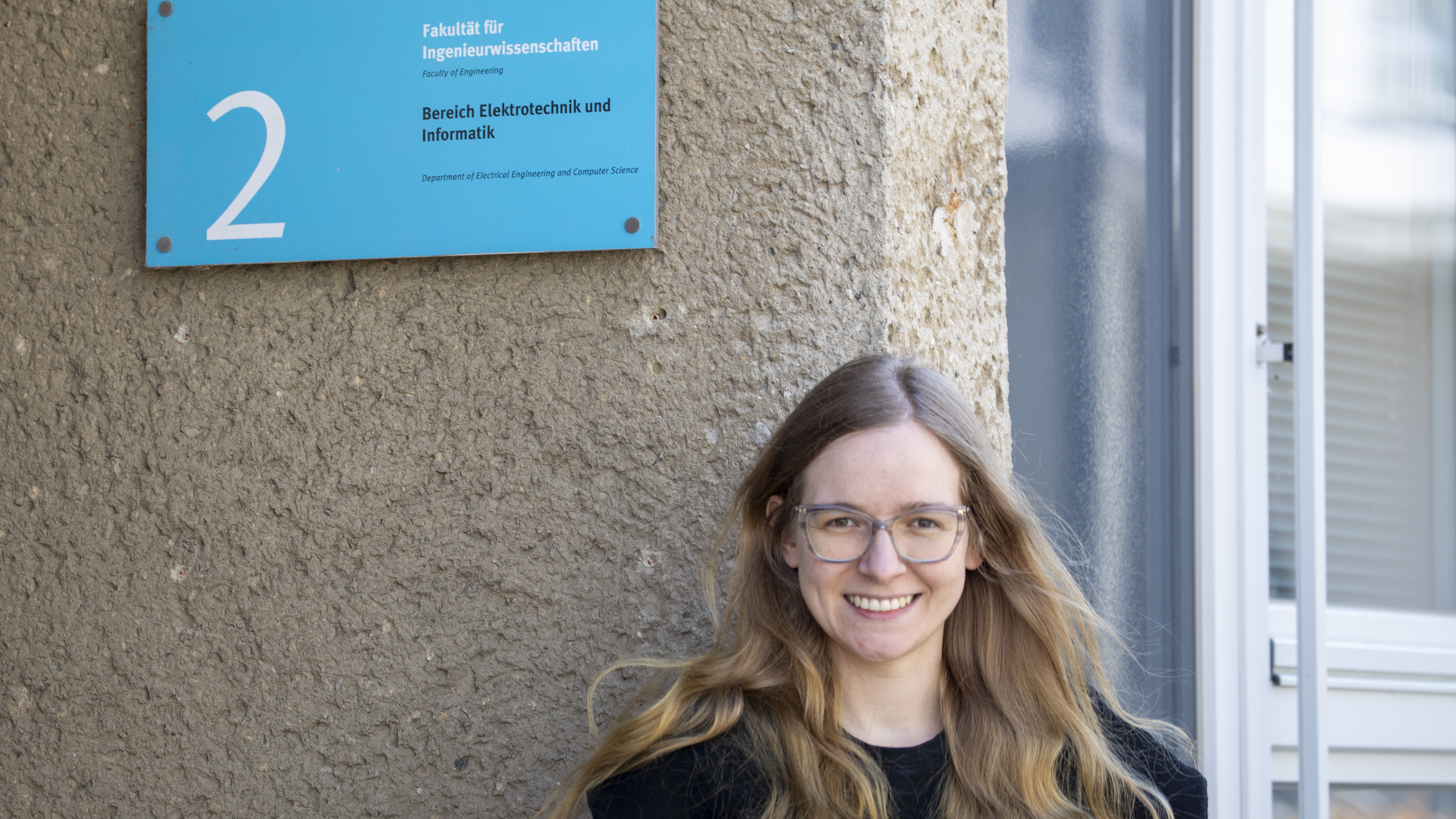 Die junge Frau hat labnge offene Haare, die im Wind wedeln.Sie steht vor einer Hauswand, an der ein blaues Schild mit dem Namen des Bereiches und der Hausnummer 2 hängt.