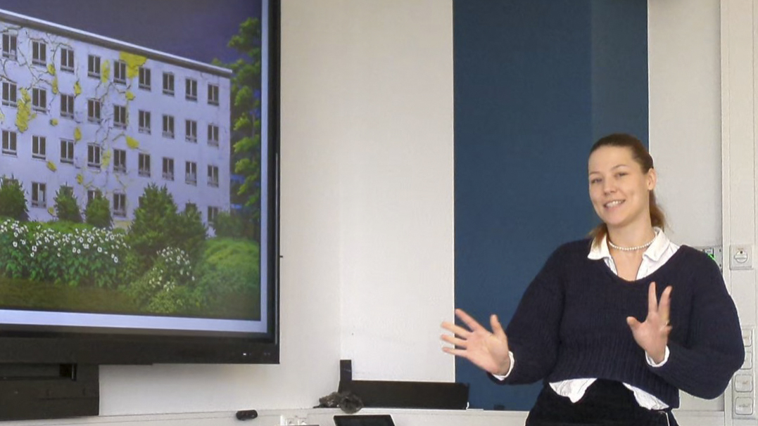 Die Studentin beschreibt mit ihren Händen eine Bewegung. Im Hintergrund ist auf einem Bildschirm eine Fotoanimation zu sehen.